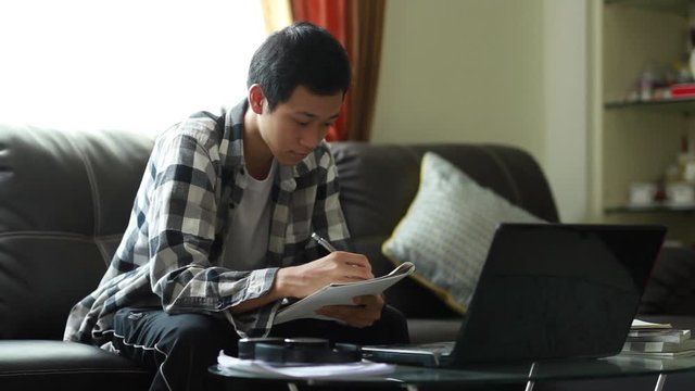 Asian teenager writing a note follow his tutor through online class by use a laptop computer at home living room.