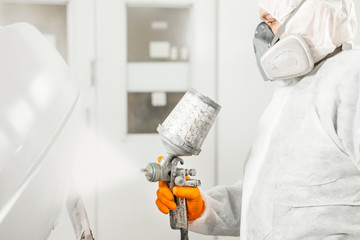Car painter spraying white paint with spray gun on vehicle in paint chamber.