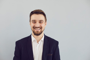 Positive bearded guy in a jacket smiling looks at the camera on a gray background.