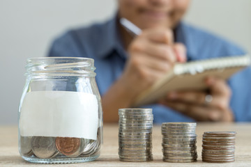 Money savings concepts glass bottle and stack coins with blur man who's taking notes about saving money for family and family wooden dolls.on wooden table with blur background and copy space.