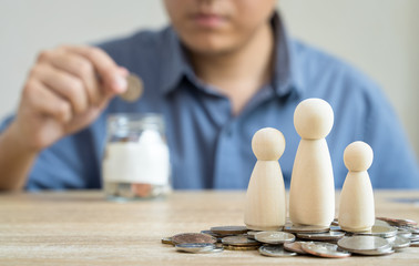 Money savings concepts Family wooden dolls on coins with blur Men are coining in Glass bottel for saving money for families with on wooden table with blur background, selective focus.