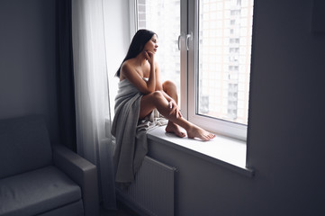 Beautiful bored slender brunette woman sitting next to window windowsill under warm grey blanket at home. Self isolation quarantine during Corona virus pandemic. COVID19 stay home save lives concept