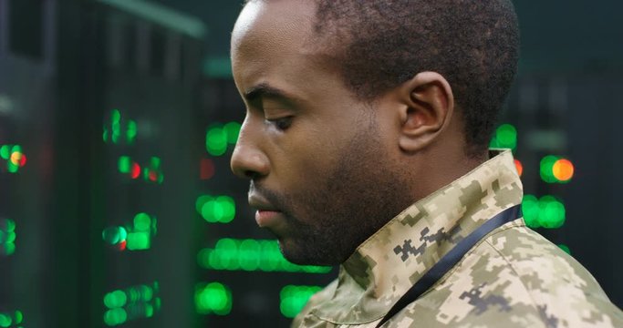 Close up of young African American army technician in camouflage unform standing at servers in secret monitoring military center and checking data. Militarian worker controlling digital facility.
