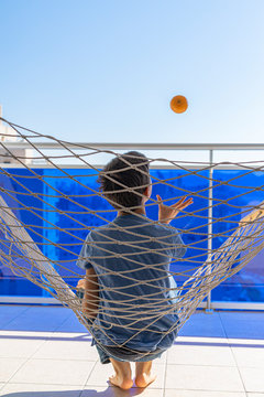 Child Playing With Orange In Hammock Hanging On Balcony Of Apartment. Back View