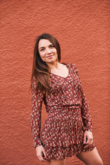 Outdoor portrait of pretty young woman with long straight brown hair, posing on terracotta color wall background, wearing mini dress