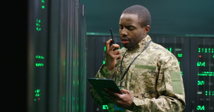 African American military man in uniform working in servers room, holding tablet device and speaking in walkie-talkie. Transmitting spying information in army. Digital data. Warfare concept.