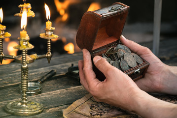 Pirate is looking on the treasure chest with ancient coins in his hands close up on burning fire...