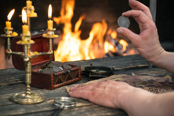 Pirate is looking on the treasure map and ancient coins in his hands close up on burning fire...