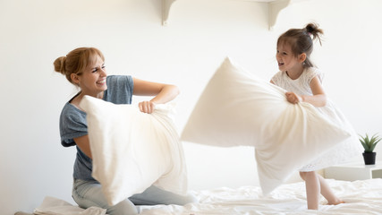 Obraz na płótnie Canvas Happy young mother pillows fight and laughing with daughter on bed at bedroom. Smiling family of mom and little cute girl having fun and playing with cushions at home together.