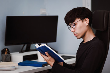 Young handsome asian man reading book at work desk late at night, Knowledge and learning concept.
