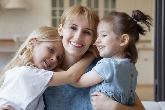 Close up headshot portrait picture of smiling mother hugging two daughter sitting on coach at home. Happy young parent with little children embracing looking at camera posing for photo in living room.