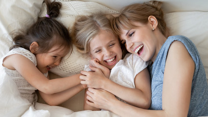 Cheerful family of smiling mother with two daughter lying down together on bed white bedding smiling looking at each other feels happy. Concept of love, offspring, close up top view.