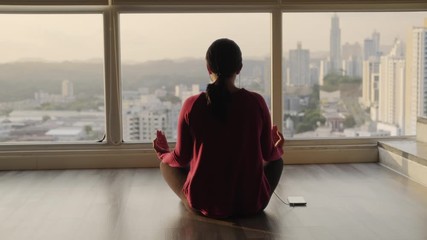 Girl doing yoga position and sitting on floor at home at sunset. Young woman relaxing and meditating with app on smartphone. People and lifestyle with mobile telephone, health, relax with cell phone