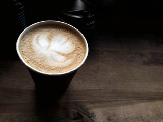 Сappuccino with drawing of a lotus flower on a foam in a black paper cup stands on a wooden table near the window with natural light. Photo cappuccino close up, top view and side view.