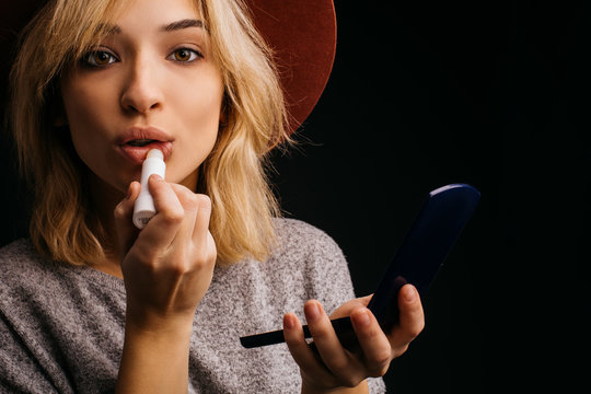 Beautiful amazing young woman using lipstick for lips. Putting some make up on face. Posing on camera. Hold fasial powder with small mirror in it. Isolated over black background.