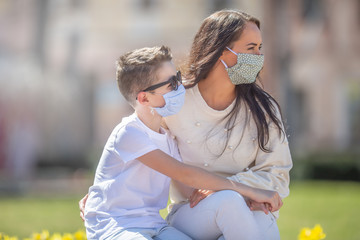 Mother and son with face masks on their faces both look to the right on a sunny day in the park