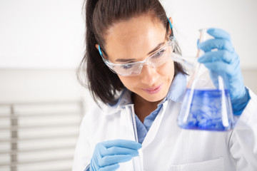 Female chemist wearing protective goggles smiles when holding a test-tube, mixing blue liquid inside the conical flask