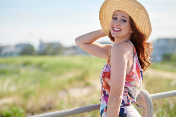 Stunning young red-haired woman in print dress and large woven hat stands on boardwalk near beach - summer vacation