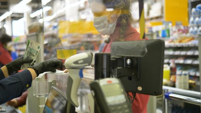 Protective measures against the spread of coronavirus. Cashier in rubber gloves gives change in paper bills to the buyer through a protective screen. Medical mask for protection from covid-19.
