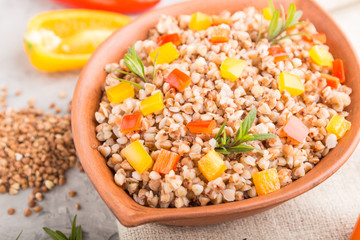 Buckwheat porridge with vegetables in clay bowl on a gray concrete background and linen textile. Side view, close up, selective focus.