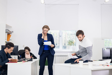 A woman talking over the phone in an open space office while reading some information from the blueprint in her hands