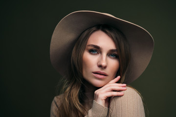 A young attractive blonde woman in a beige sweater, fedora hat and brown leather trousers poses on a dark green olive background. Studio portrait of a beautiful girl.