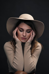 A young sensual sexy woman with a beautiful face and clear natural skin poses in the studio against a black background. Beige slinky sweater and hat with fields.