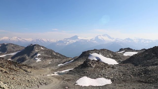 Summer day on the top of the mountain, people hiking, jogging between left over snow patches. Endless range of mountain peaks in the background. 