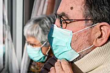 senior couple, with protective face masks, at home looking through the window. Concept of coronavirus quarantine stay home and social distancing. Lockdown people. elderly and retired lifestyle .