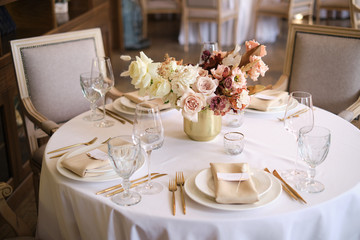 Luxury cozy autumn wedding table decoration in the restaurant. Fresh and dried flowers, roses, carnations. Beautiful table setting: golden appliances, fork and knife, calligraphy guest seating card.