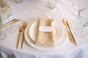 Luxury cozy autumn wedding table decoration in the restaurant. Fresh and dried flowers, roses, carnations. Beautiful table setting: golden appliances, fork and knife, calligraphy guest seating card.