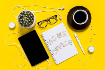 Flat lay of modern workspace with tablet, earphones, coffee cup, notepad, pen, eyeglasses and cactus. Letters saying Home Office. Top view on yellow background