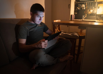A man at home sitting on the couch at night and uses the tablet to search for work, news,...