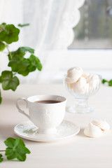 Black tea in a white porcelain Cup on a saucer, meringue in a sweet bowl and next to the Cup on a white wooden table. In the background is a window with a curtain