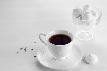 Black tea in a white porcelain Cup on a saucer, dry tea scattered on the left, meringue in a sweet vase and next to the Cup on a white wooden table