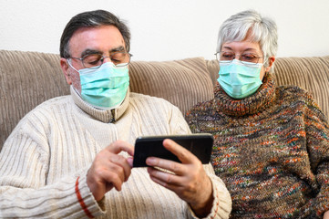 Coronavirus. Stay at home, lifestyle.Cheerful Elderly couple sitting on a sofa on a quarantine at home, making a video call with the smartphone. Senior Couple wearing protective masks .
