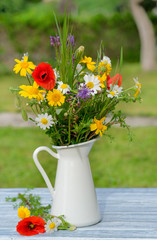 Wild flower bouquet in white jug on blue vintage wooden table. Still life with bouquet of spring wildflowers. Spring, summer day. Beautiful floral background. Cute vertical card.