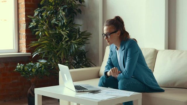 Young female in blue suit smiling and clenching fists then making phone call to share success after finishing remote project on laptop at home