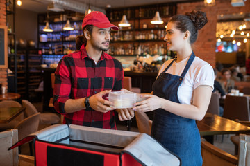 Young waitress in workwear passing plastic container with fresh food to courier