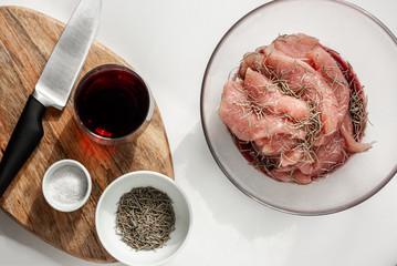 Turkey in a red wine, rosemary and salt marinade in a glass bowl on a white background. Nearby is a wooden cutting board and marinade ingredients. Cooking concept