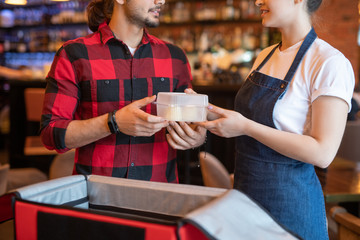 Young courier in red flannel taking container with fresh food ordered by client