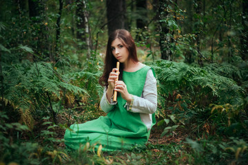 A medieval woman in a green dress plays on a wooden flute sitting in fern bushes. A girl in a gloomy forest plays music on a handmade pipe. Young witch in a twilight fantasy forest. © Михаил Гута