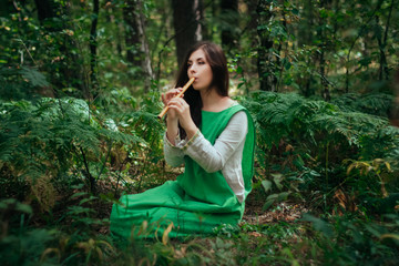 A medieval woman in a green dress plays on a wooden flute sitting in fern bushes. A girl in a...