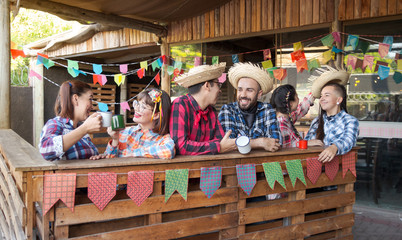 Brazilian June Party, typical celebration in Brazil. Joyful men and women in typical plaid shirt and straw hat having fun, drinking and talking at Arraial Party. Rustic wooden decor with flags.