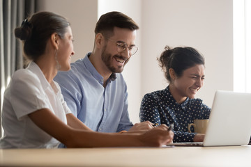 Three millennial multi-ethnic colleagues sitting at workplace feels overjoyed laughing during...