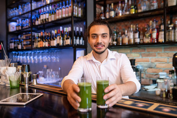 Happy young waiter or barman in white shirt passing you two glasses of smoothie