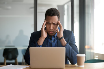 African businessman looking at laptop screen feels frustrated received bad news by e-mail, worker suffers from headache massaging temples to relieve painful feelings, pc overuse stressful work concept