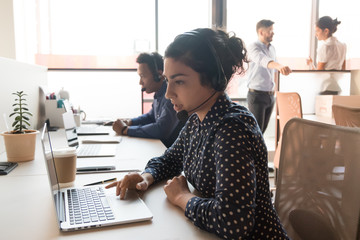 Attentive call centre indian woman employee sitting in modern shared office working using computer...