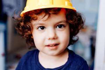 little curly boy in a construction helmet in his room on a blurry background close up