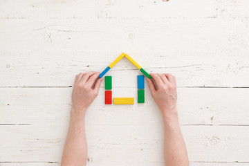 Directly above on wooden colorful bricks on a white wooden background. Hands of a man protect a house assembled from multi-colored wooden blocks. Home, family and education concept.